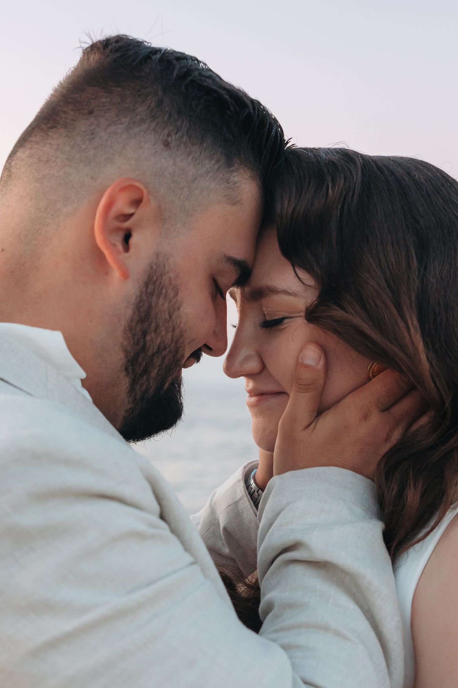 A romantic couple touching foreheads tenderly, man gently cupping woman's face, sharing an intimate moment outdoors at dusk.