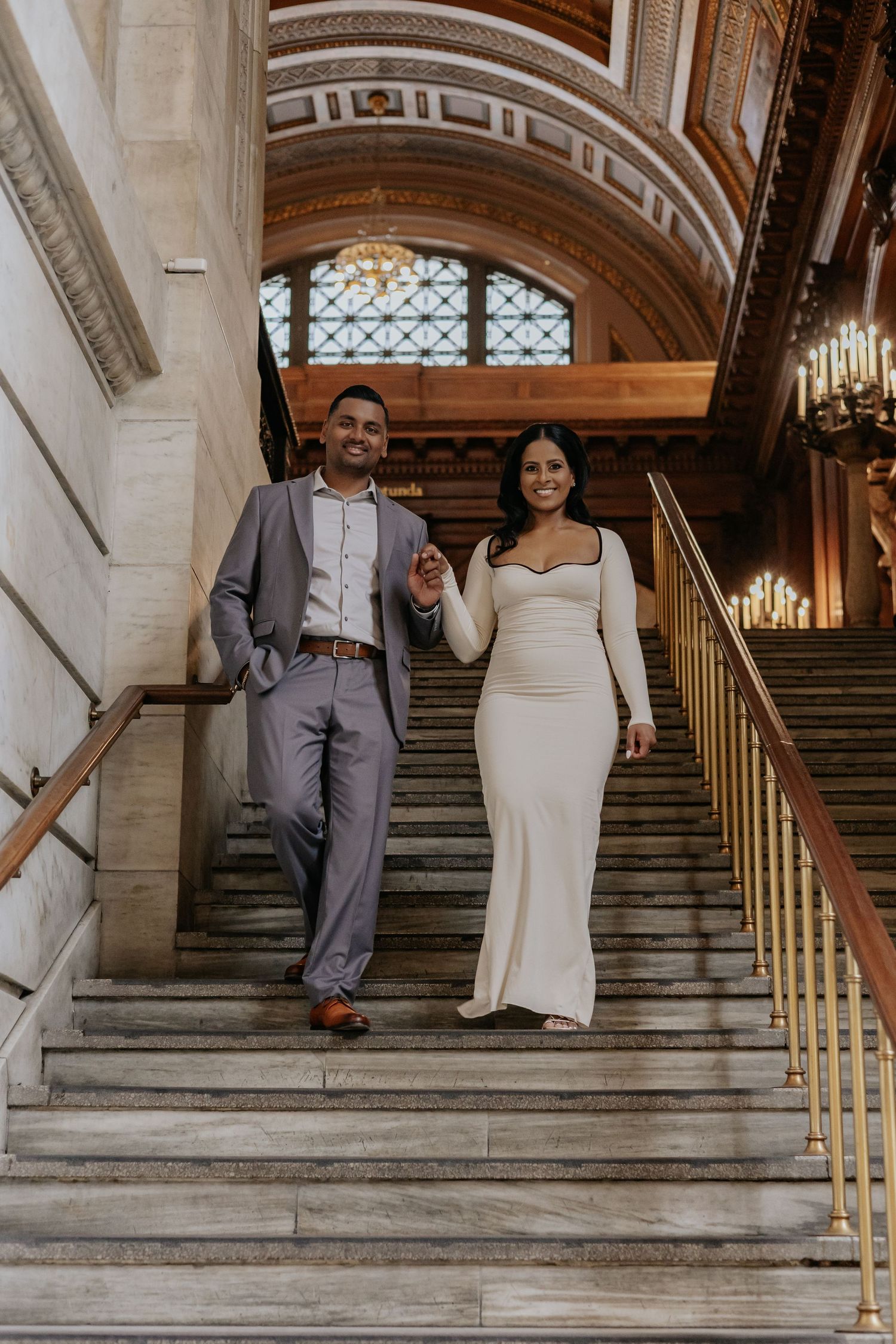 Elegant couple in wedding attire walking hand-in-hand down grand marble staircase in ornate historic building.
