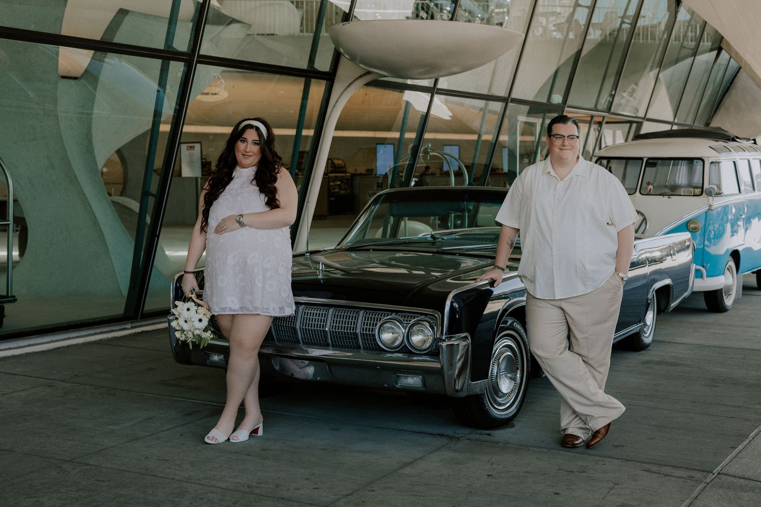 Bride and groom lean against vintage black Lincoln car outside iconic TWA Hotel terminal.