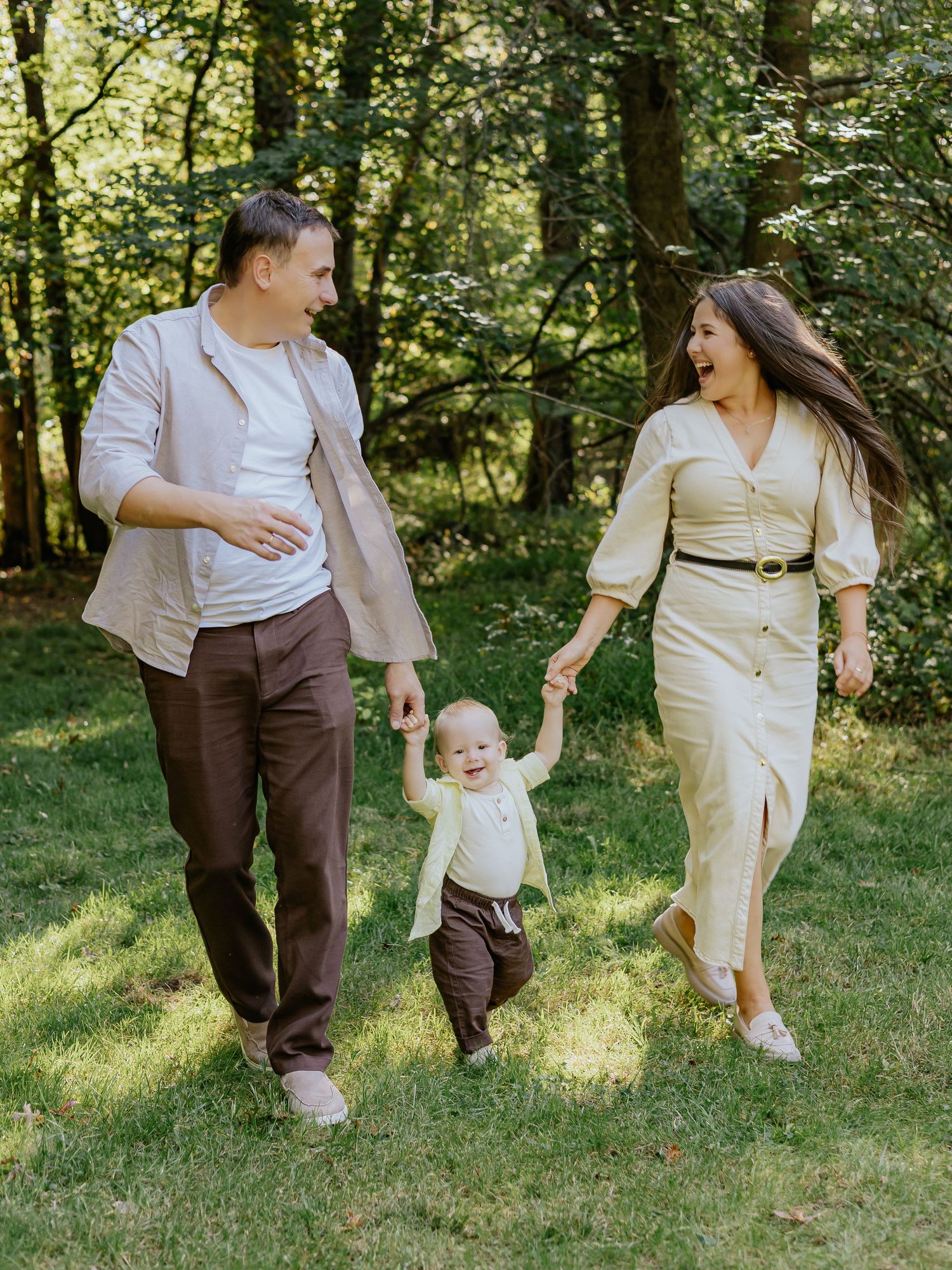 Happy young family with toddler walking in sunny green park, parents holding child's hands outdoors.