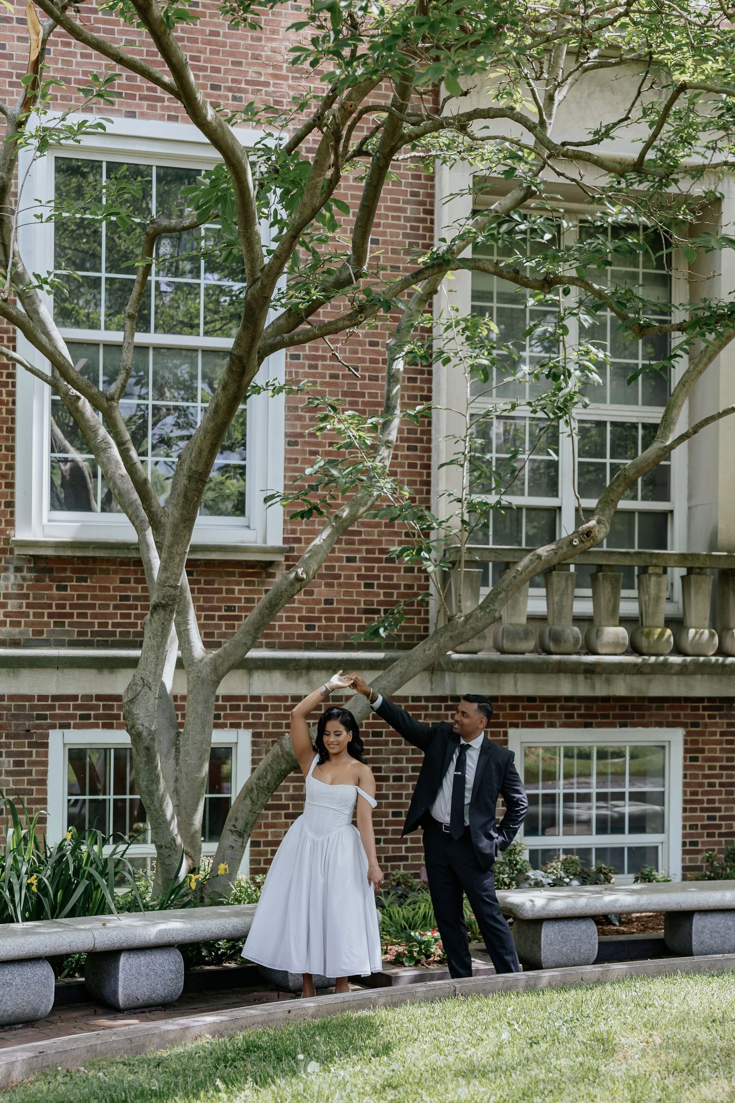 Bride twirls under groom's hand in front of a brick building surrounded by trees.