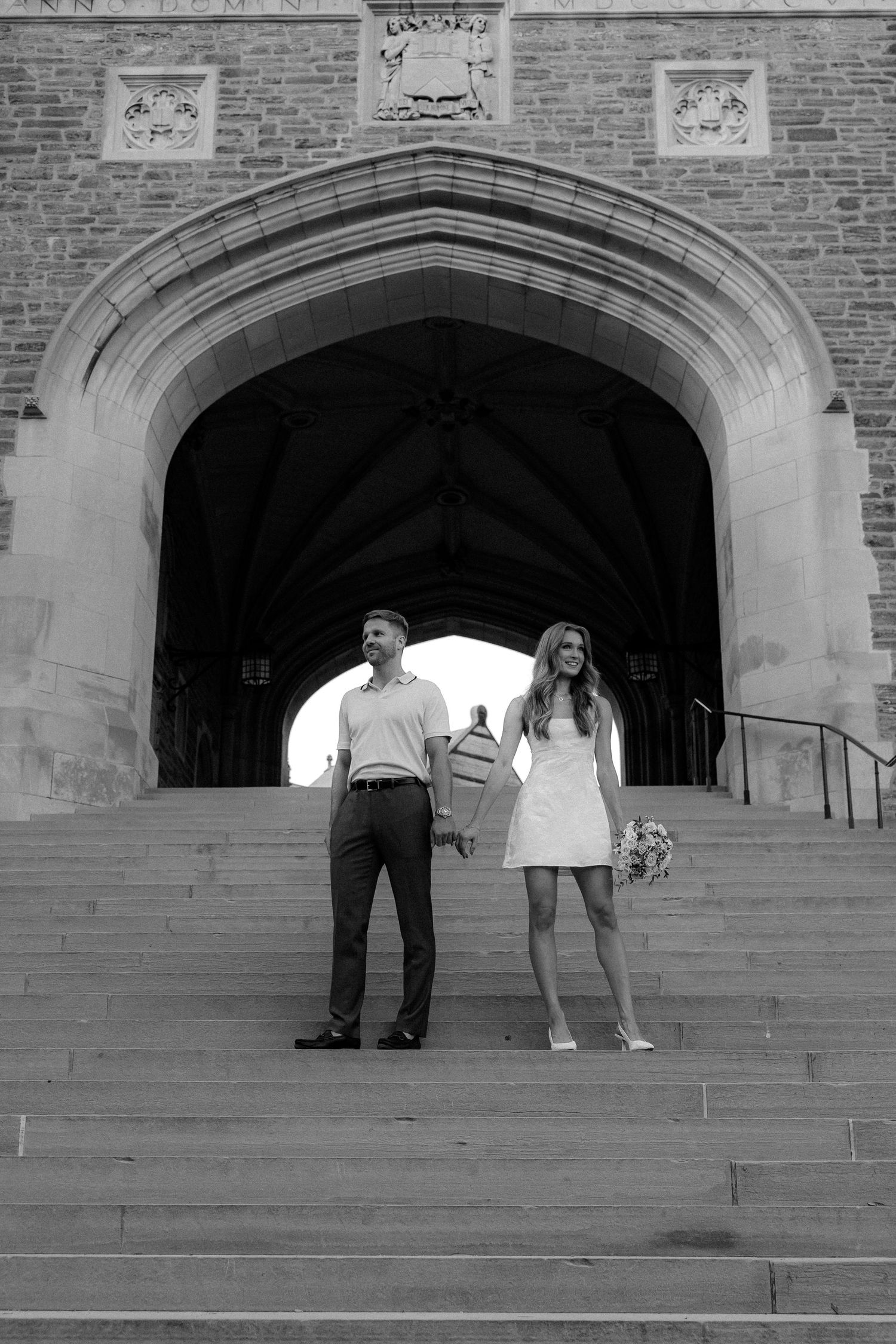 Black and white photo of a couple holding hands on stone steps beneath a grand Gothic arch at a university.