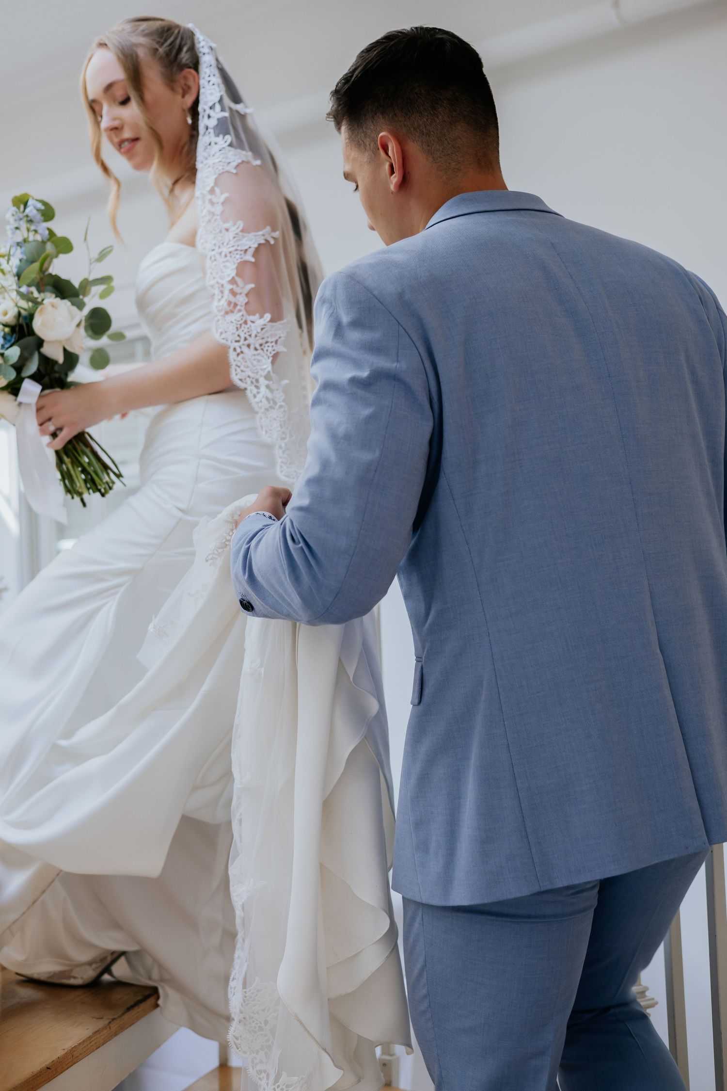 Groom in blue suit helping bride in lace veil carry her wedding dress up a staircase.