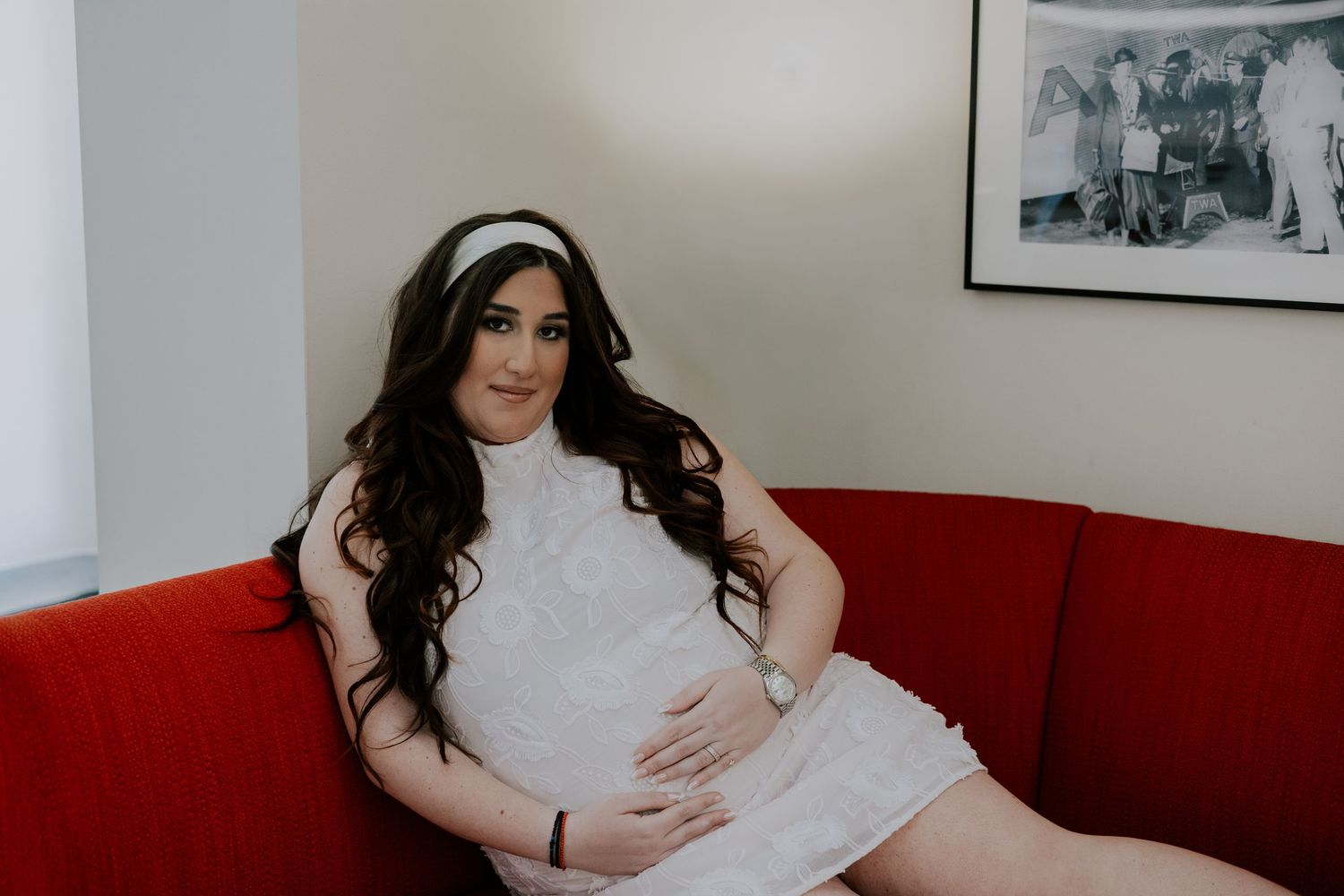 Pregnant bride in white lace dress and headband sits on red sofa holding baby bump.