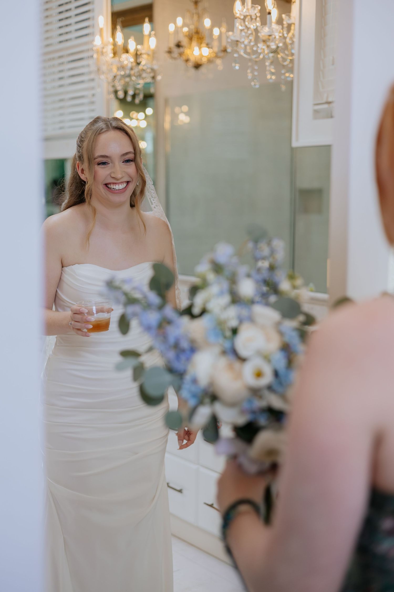 Joyful bride in strapless white gown holding drink near blue and white floral bouquet.