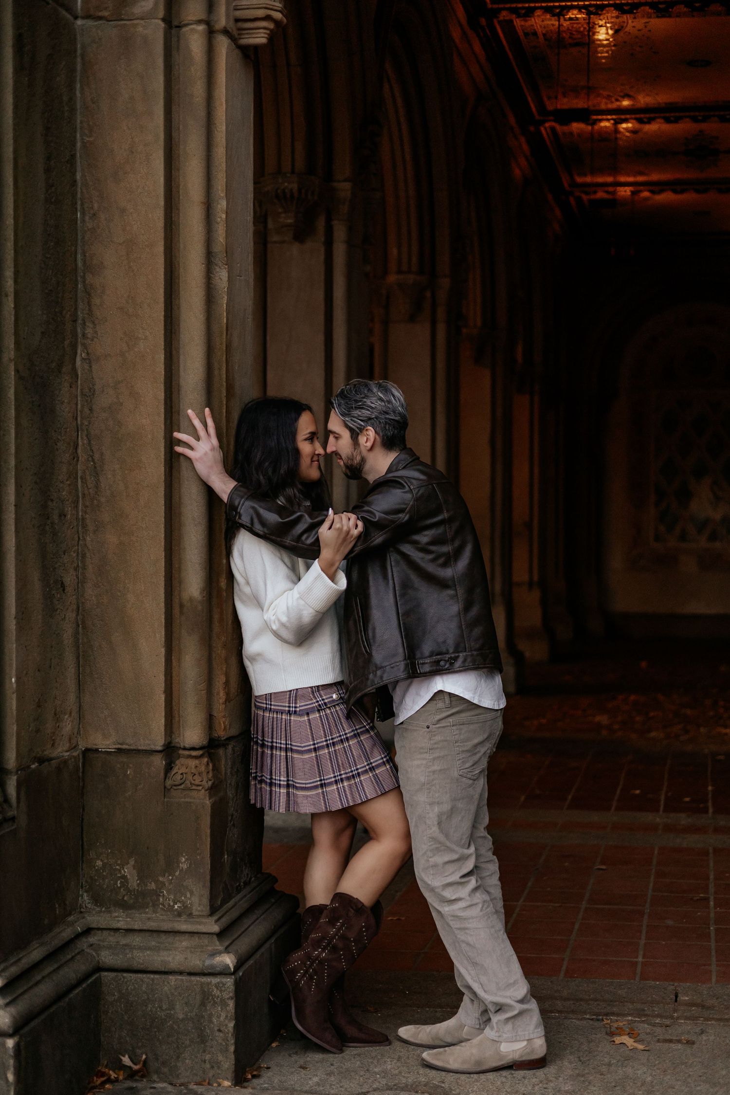 Couple sharing an intimate moment leaning against a stone column in a dimly lit archway.
