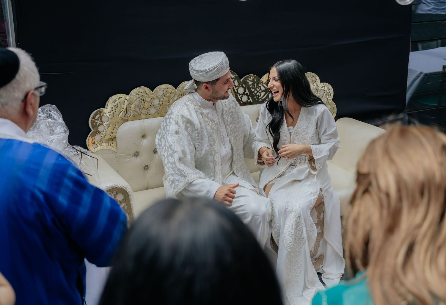 Couple in white traditional Moroccan wedding attire share a joyful moment seated on ornate gold sofa with guests watching.