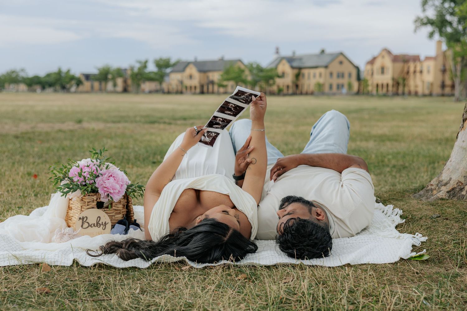 Pregnant couple lying on white blanket in park, holding ultrasound photos, with flowers and baby sign nearby.