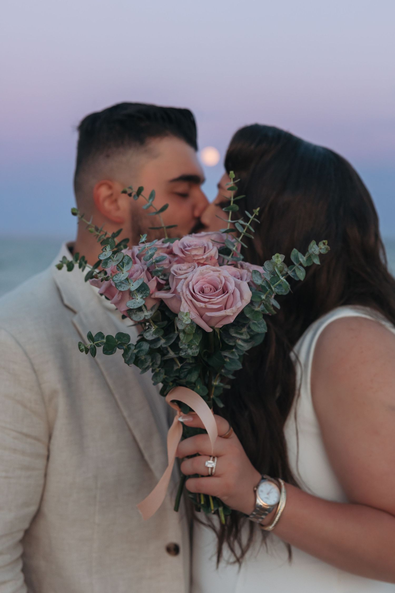 Couple sharing a romantic kiss behind a bouquet of pink roses and eucalyptus at twilight on a beach.