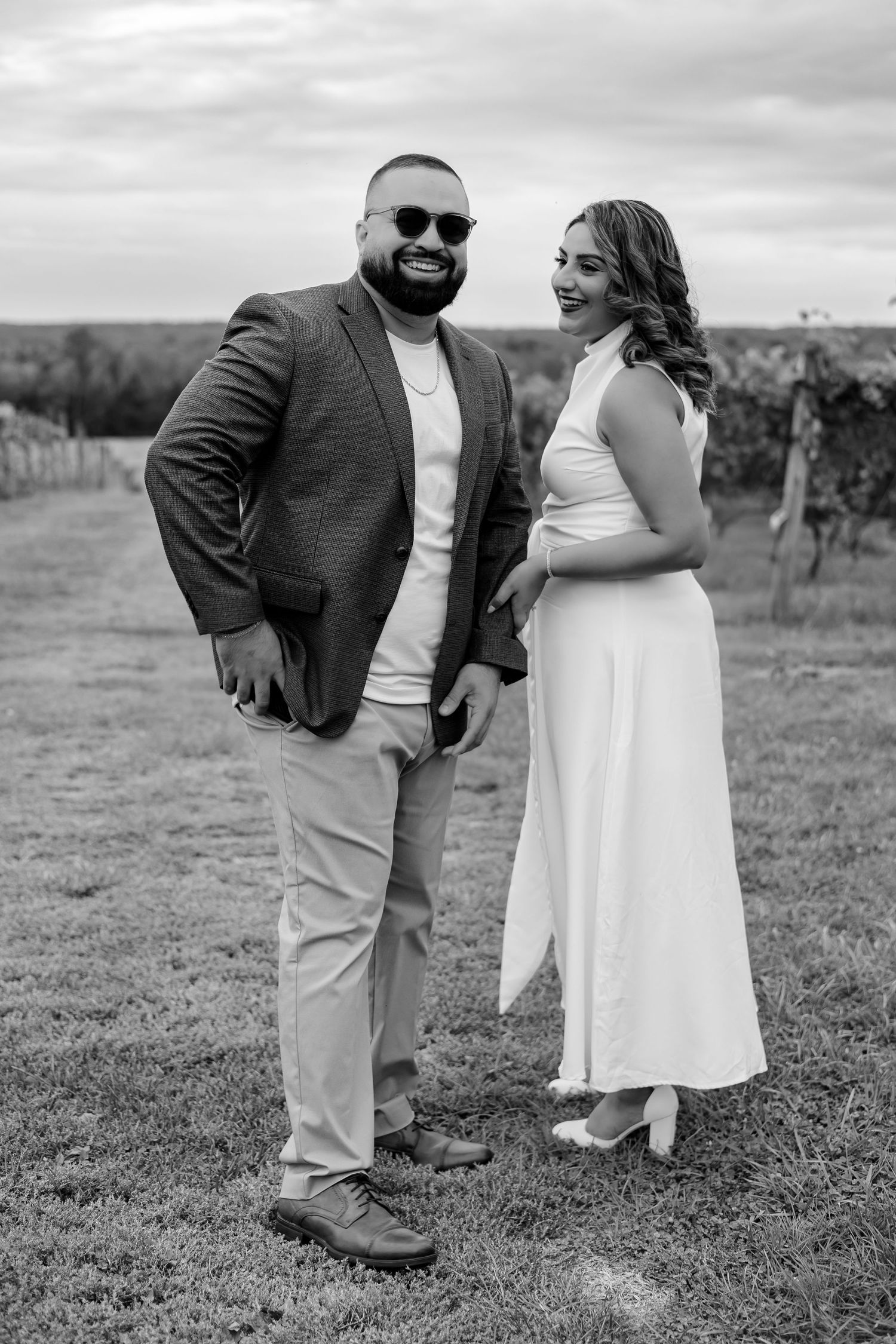 Black and white portrait of smiling couple in elegant attire standing in vineyard under cloudy sky.