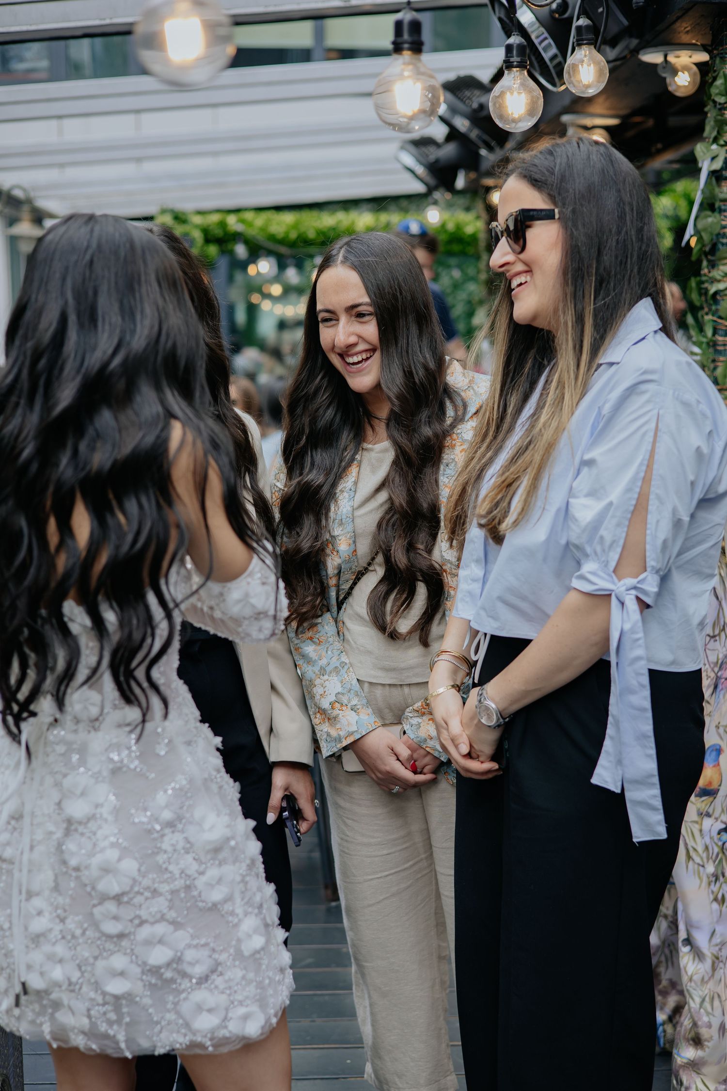 Three elegantly dressed women laughing together at an outdoor wedding reception event.