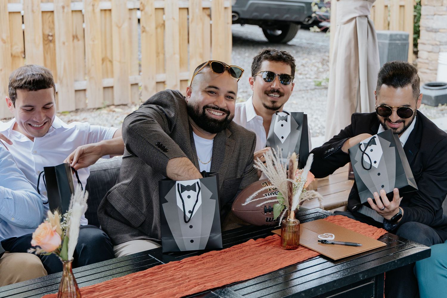 Four smiling men holding groomsmen proposal gifts with tuxedo bags at outdoor table.