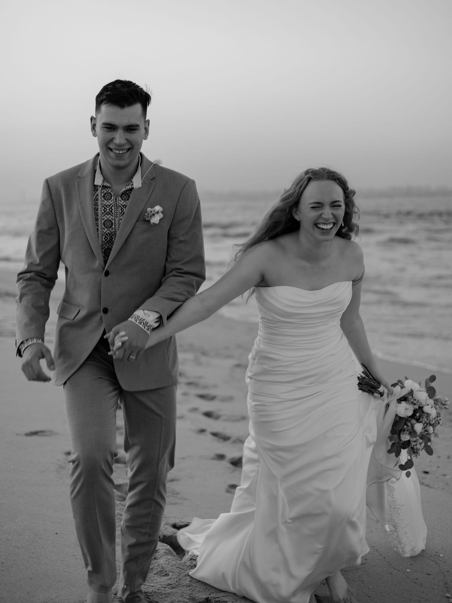 Black and white photo of laughing newlyweds walking barefoot on beach in wedding attire.