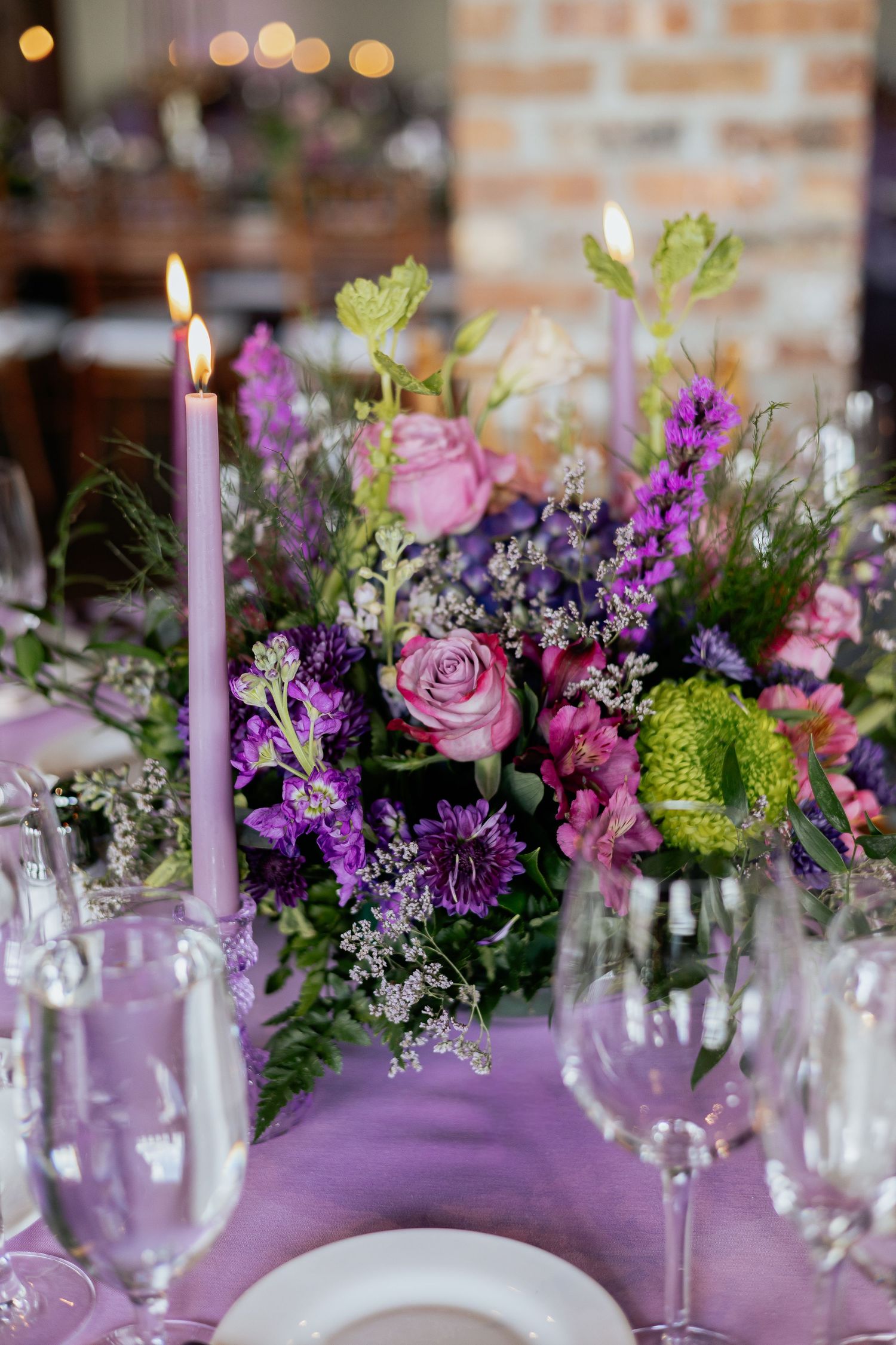 Lush purple and pink floral centerpiece with lit lavender taper candles on a lilac tablecloth at a wedding reception.