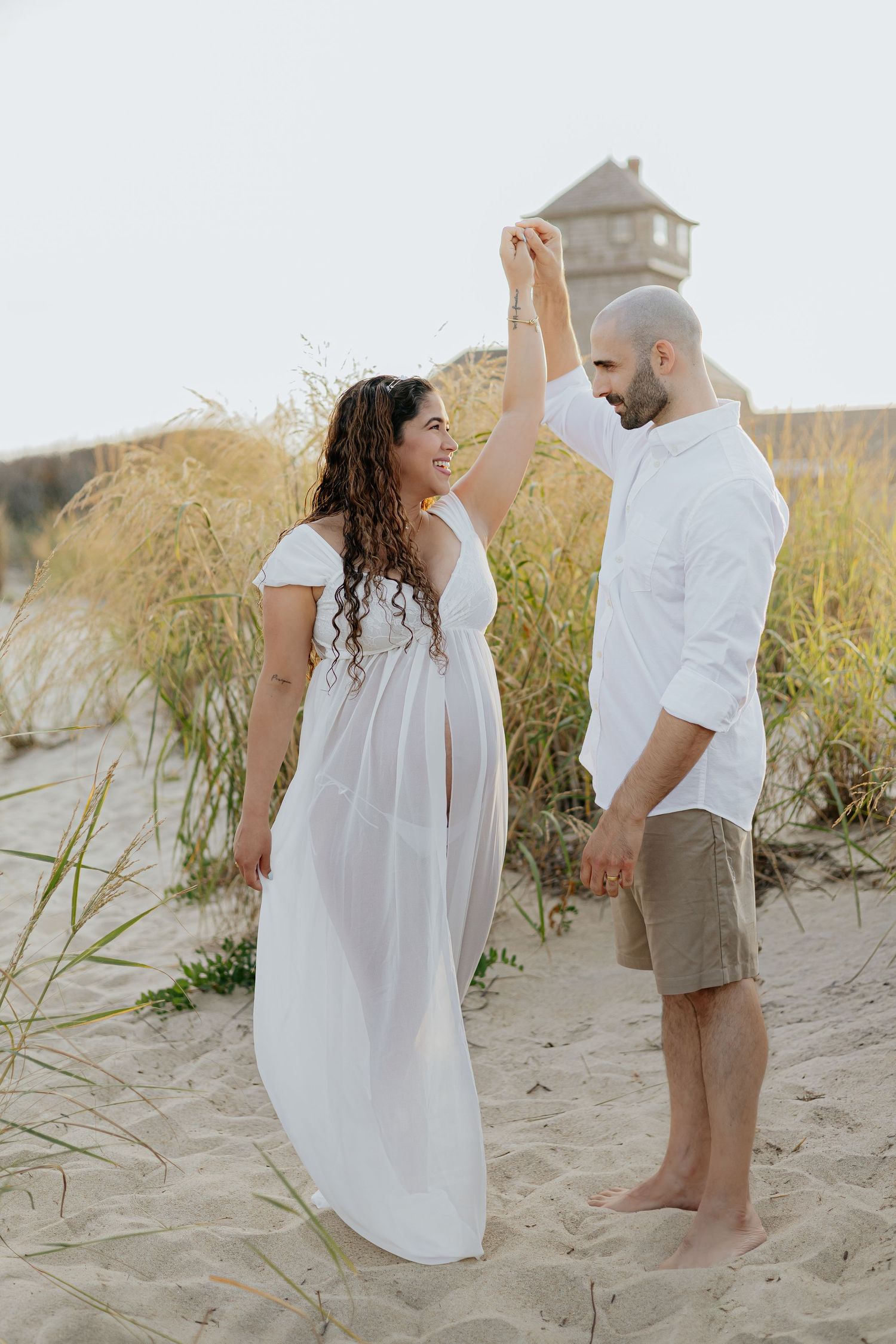 Couple dancing on beach dunes, man spinning pregnant woman in white dress during maternity session.