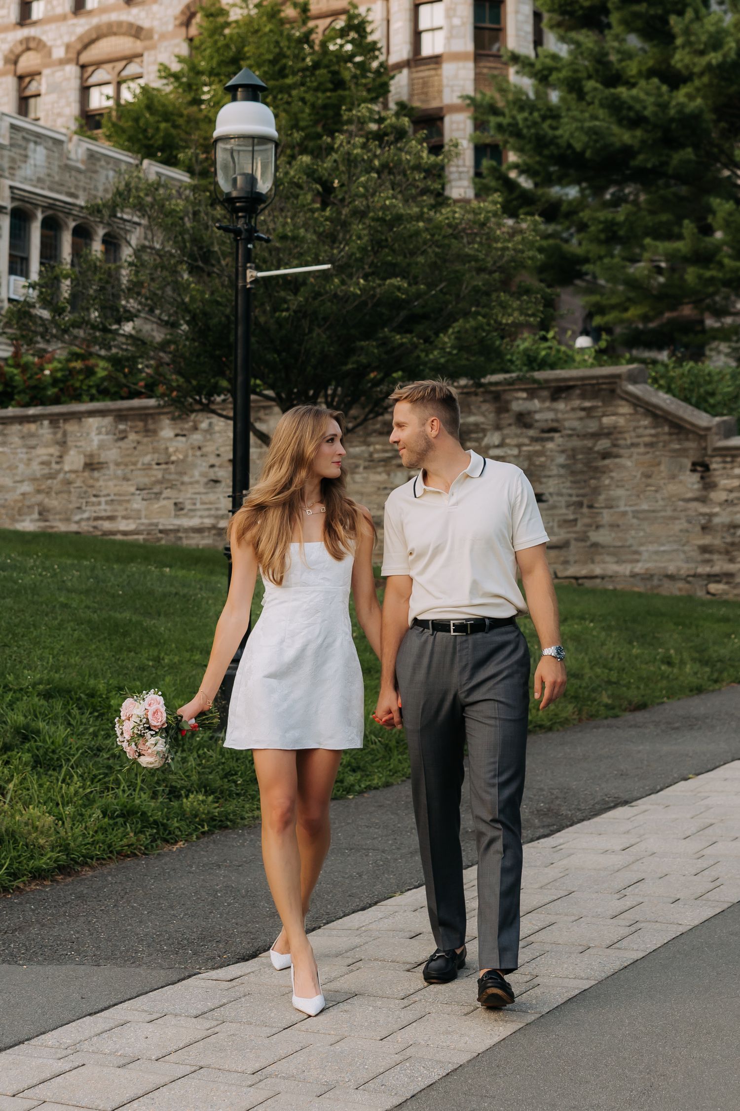 Newlywed couple walking hand in hand outdoors, bride in white mini dress holding bouquet.