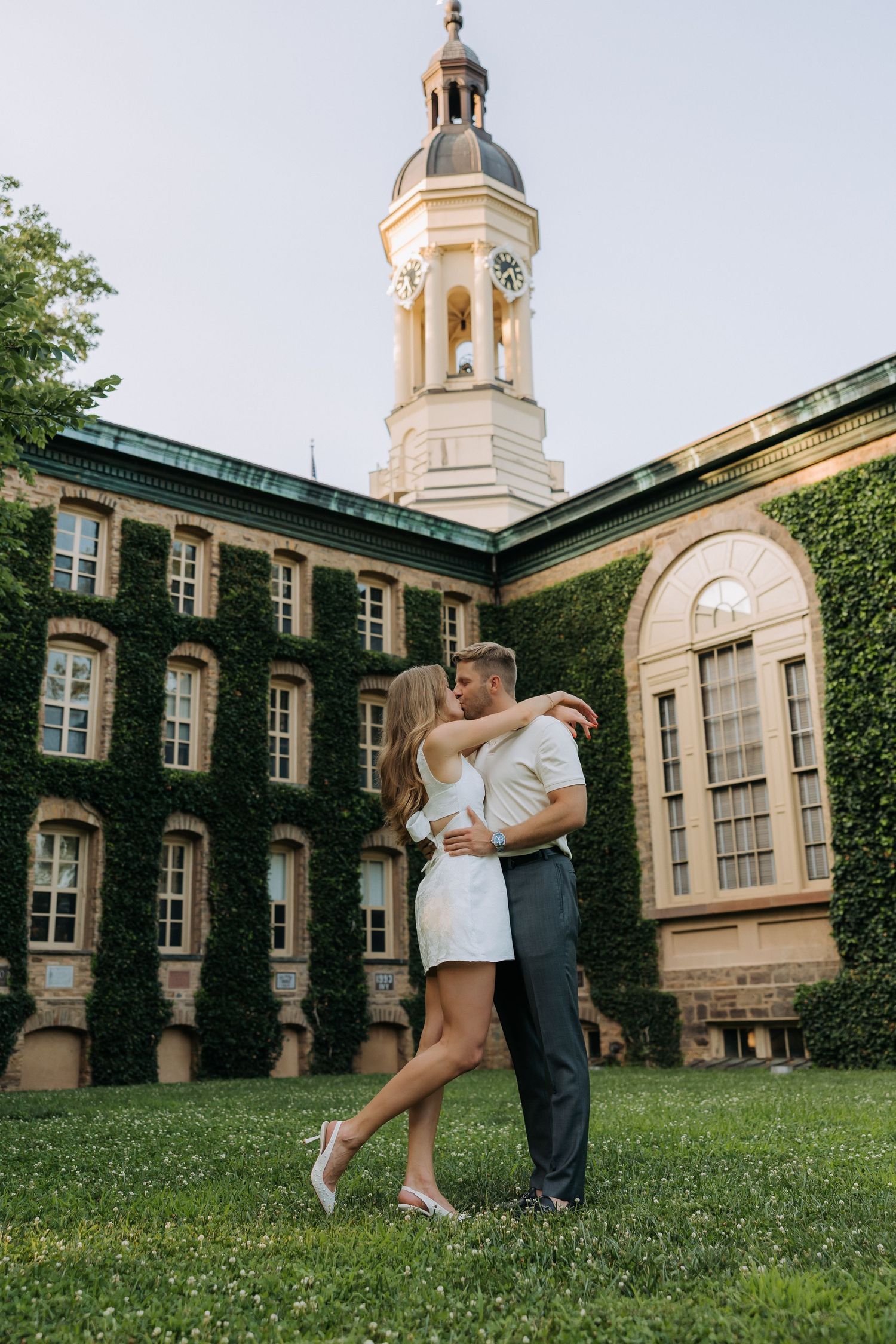 Couple embracing on green lawn before ivy-covered historic building with clock tower at dusk.