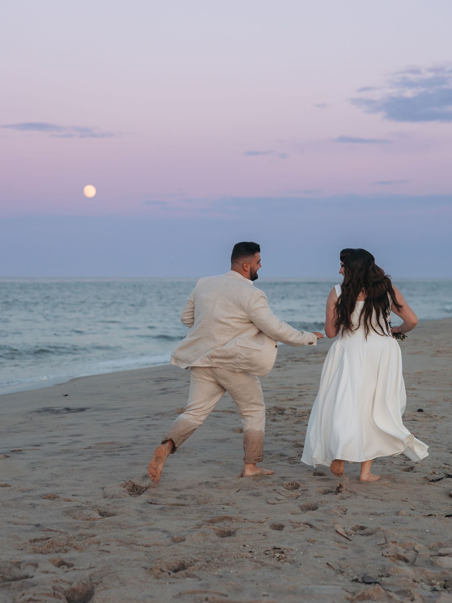 Couple in white wedding attire walking hand-in-hand on beach at sunset with full moon rising over ocean.