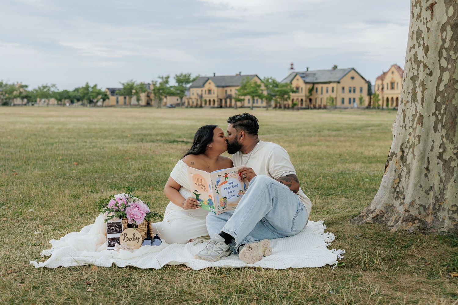 Couple sharing a kiss during a romantic outdoor picnic on a white blanket near a large tree with buildings in the background.