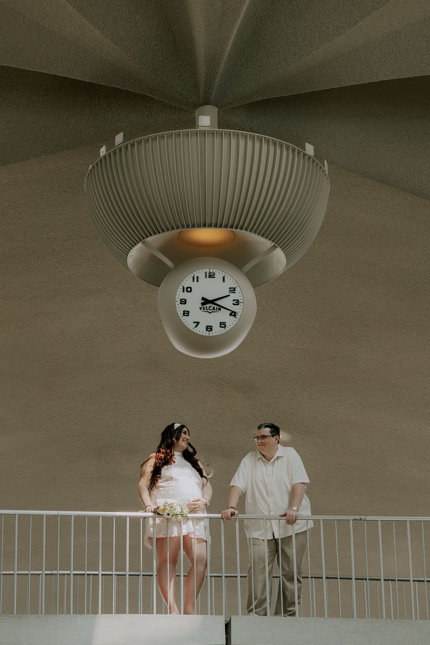 Bride and groom share a romantic moment on balcony beneath iconic TWA Hotel clock pendant.