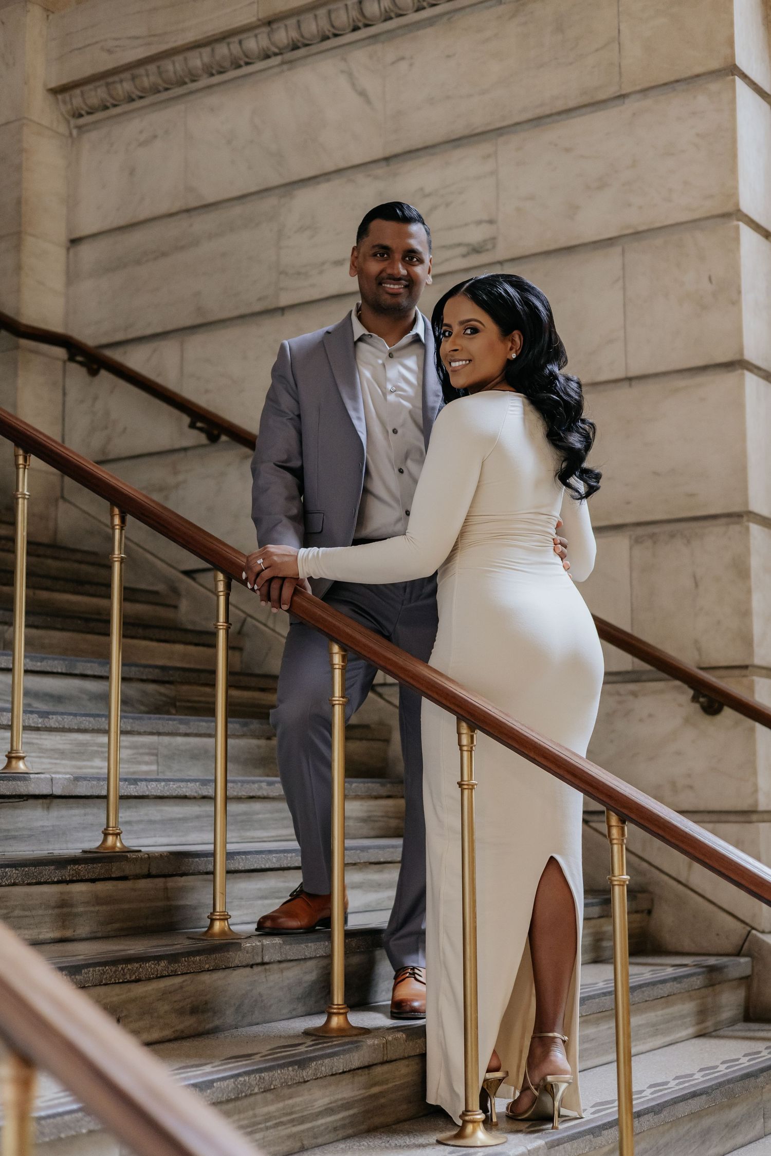 Couple posing on grand marble staircase, woman in fitted white dress smiling.
