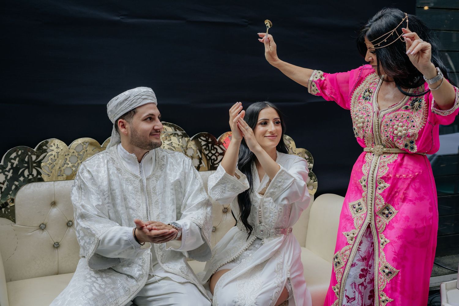 Woman in pink kaftan dances joyfully beside couple in white Moroccan wedding attire seated on ornate gold sofa.