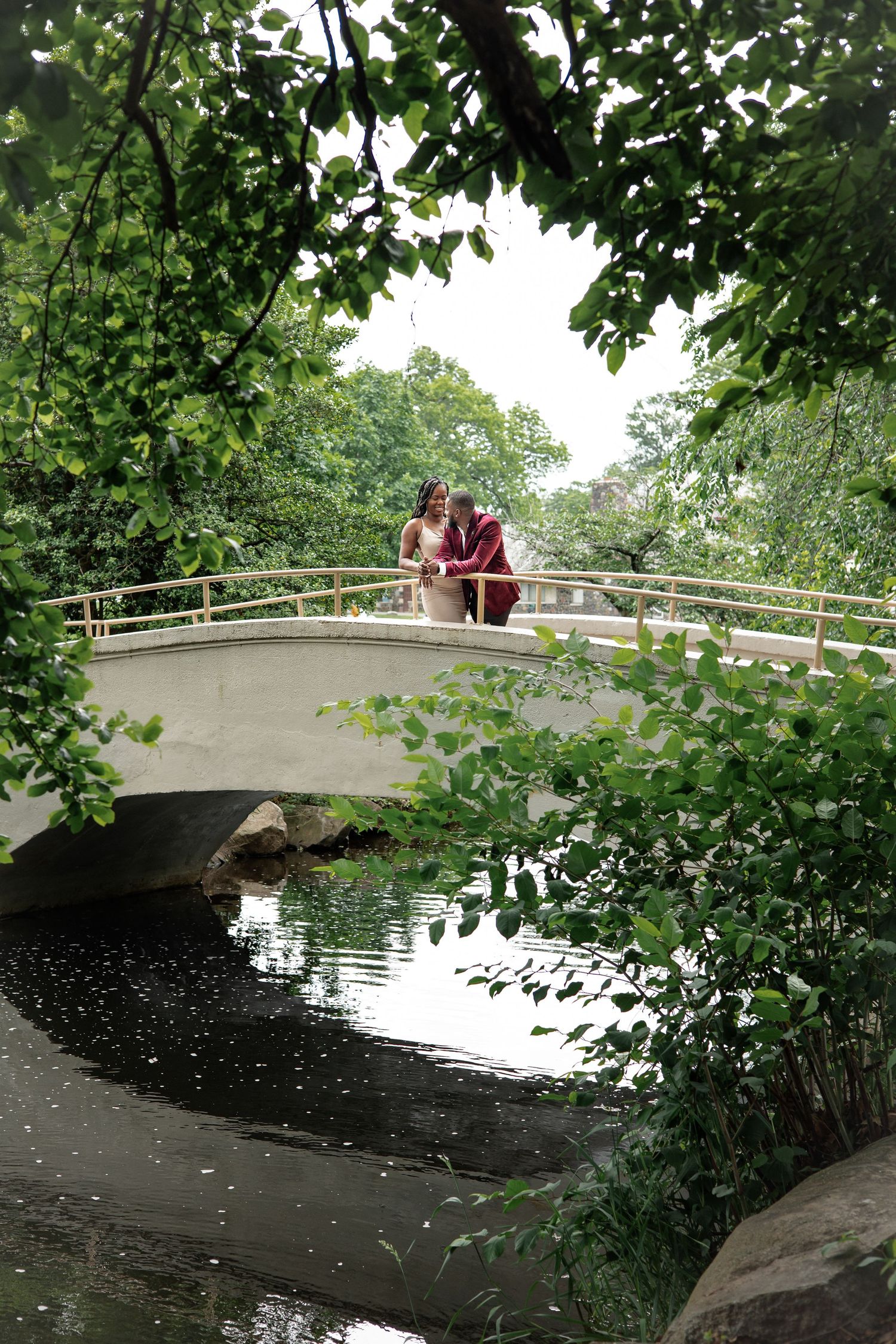 Engaged couple sharing romantic moment on stone arch bridge surrounded by lush green trees over reflecting pond.