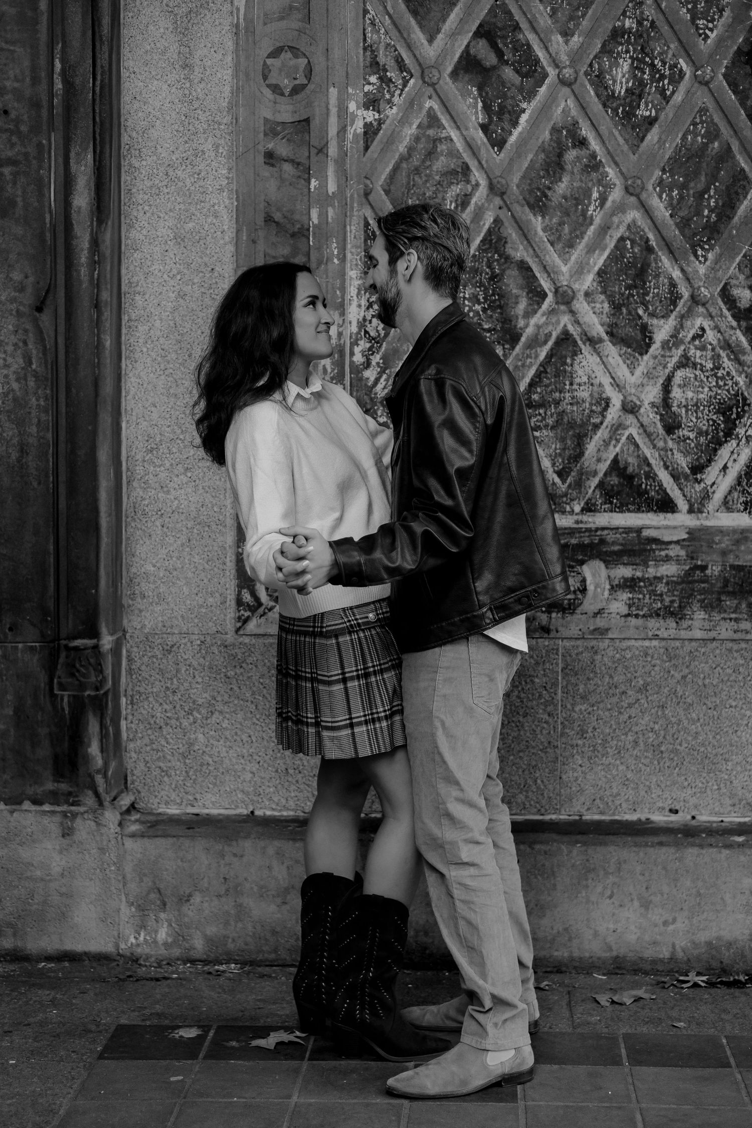 Black and white photo of a romantic couple gazing at each other near an ornate vintage wall.