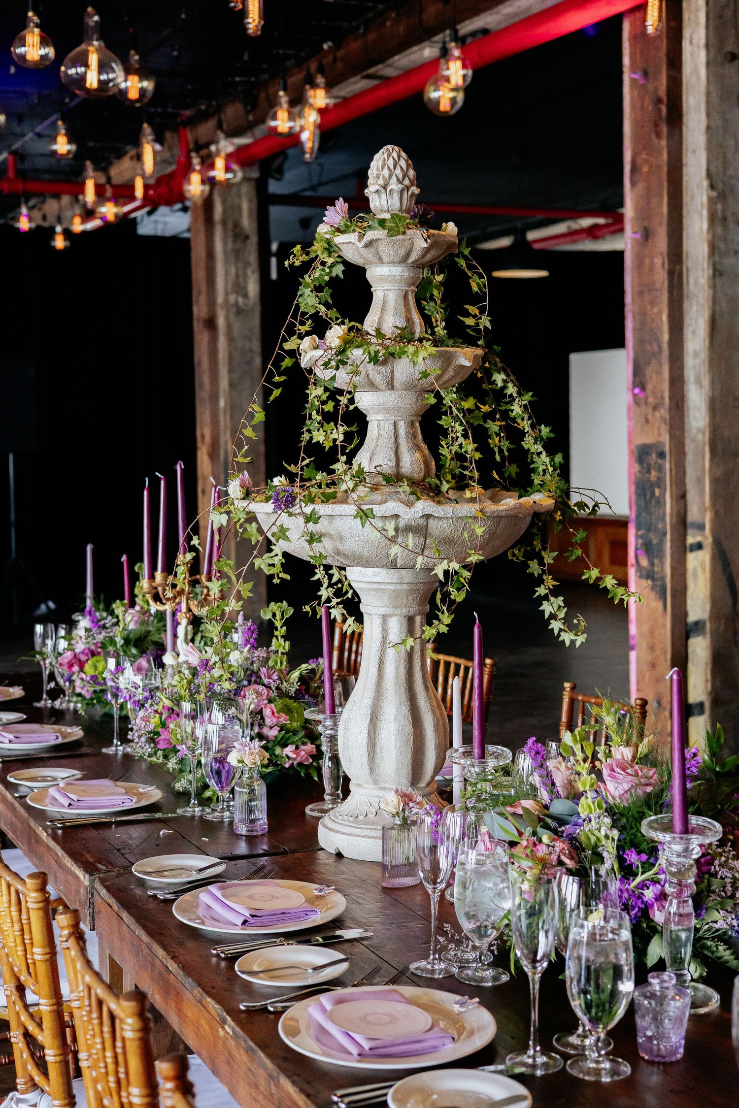 Rustic wedding reception long table with stone fountain centerpiece draped in greenery and purple taper candles.