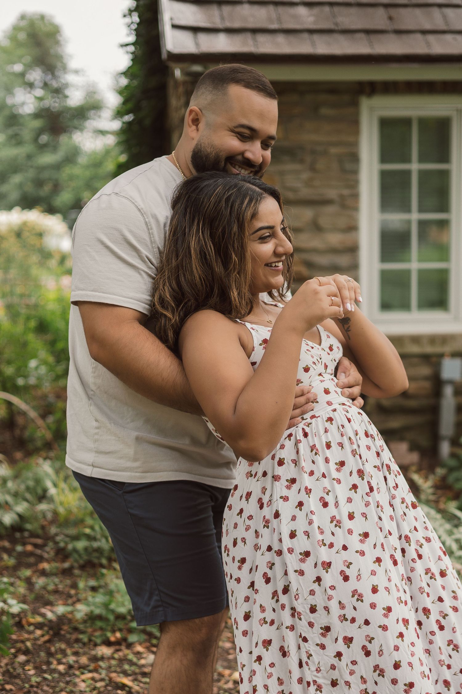 Man hugging woman from behind as she laughs in a charming garden beside a stone building.