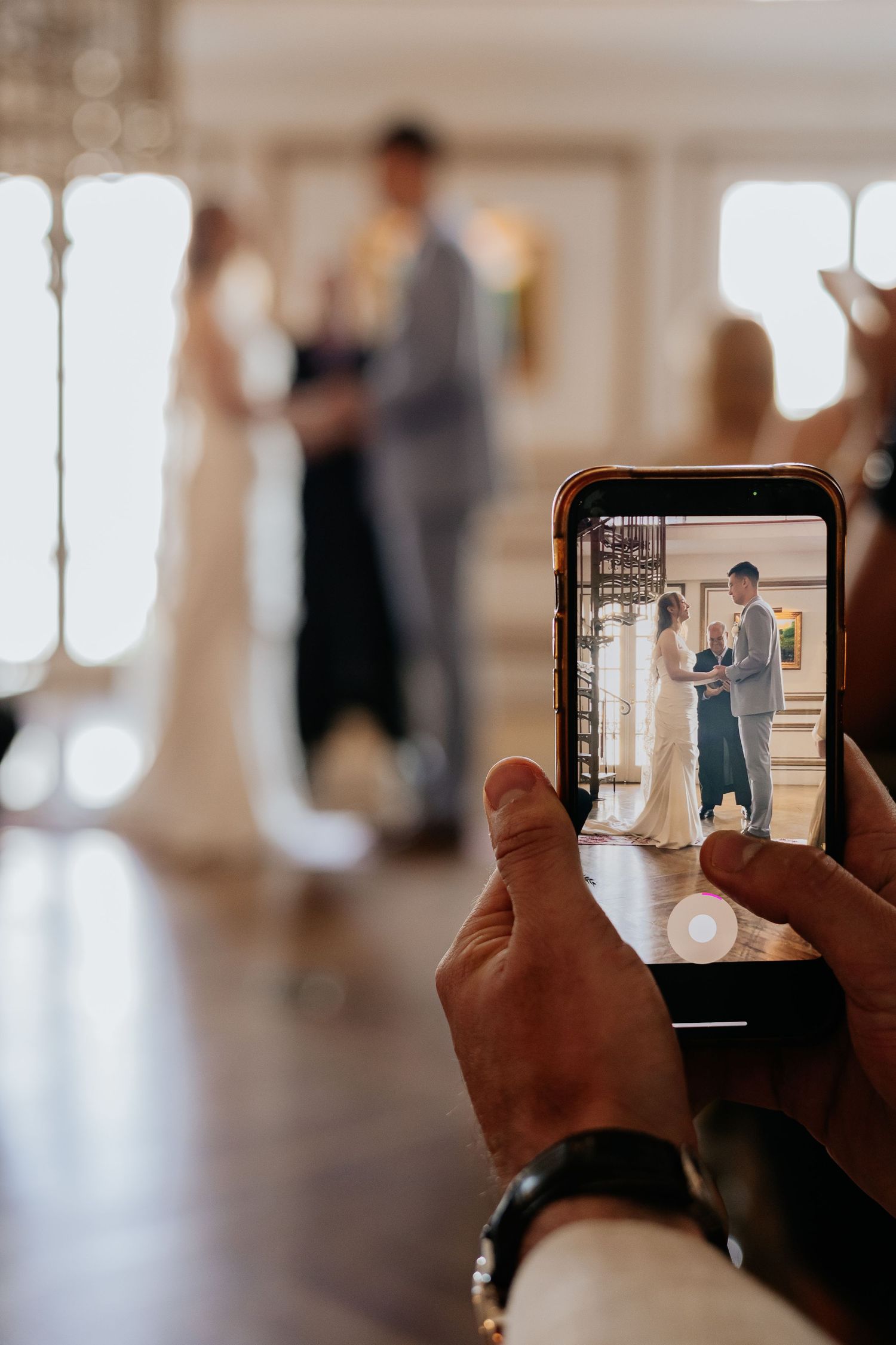 Guest photographs bride and groom exchanging vows on smartphone during elegant indoor wedding ceremony.