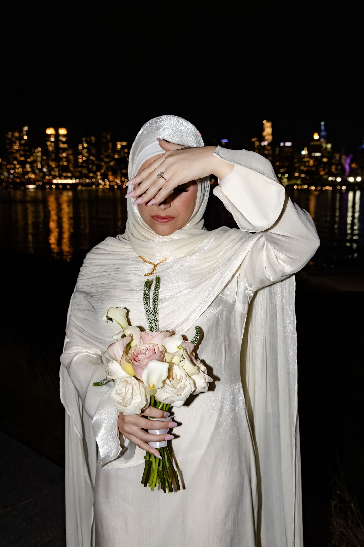 Muslim bride in white hijab covering face with hand while holding bouquet by city skyline at night.