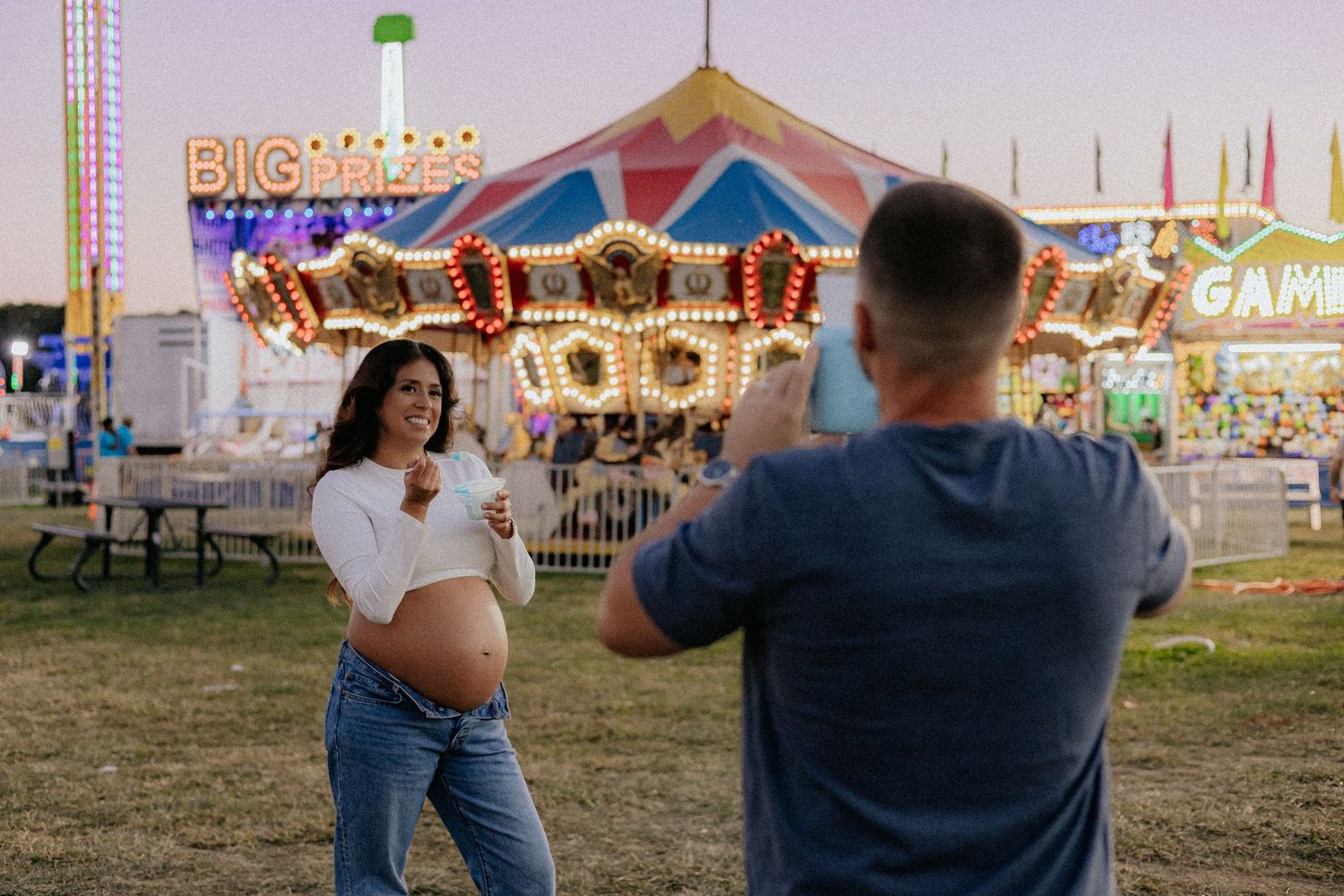 Man photographing pregnant woman posing in front of lit carousel.