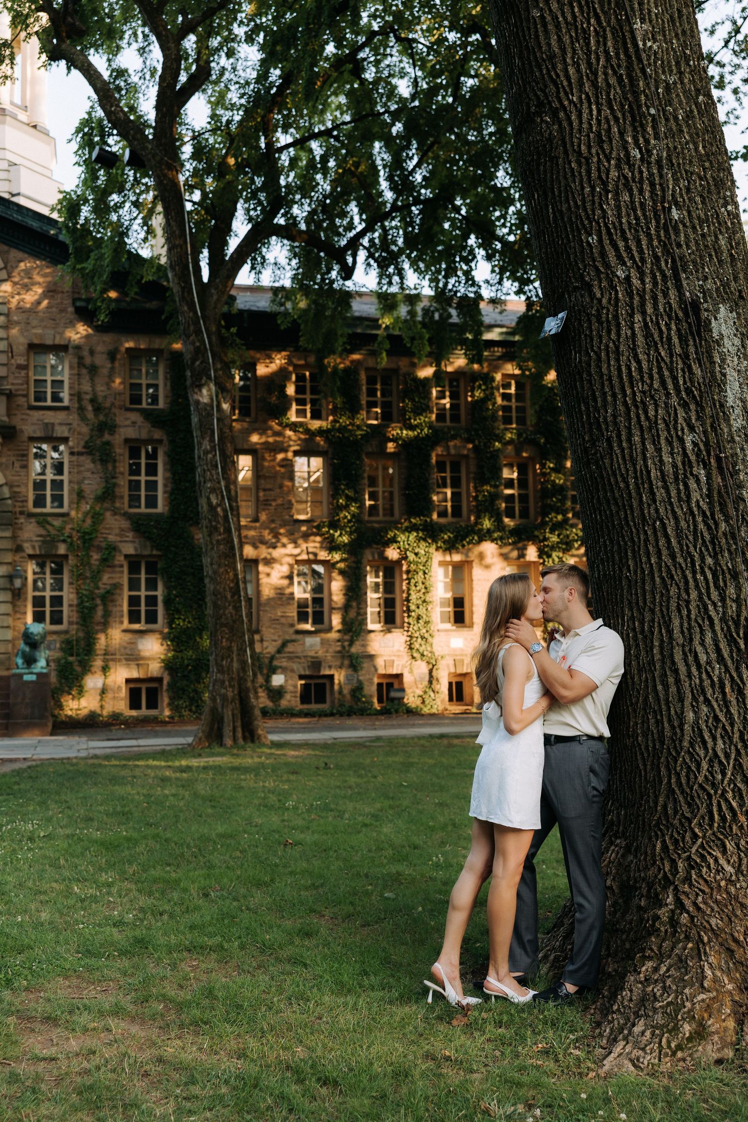 Romantic couple sharing a kiss under a large tree beside a historic ivy-covered brick building on a green lawn.