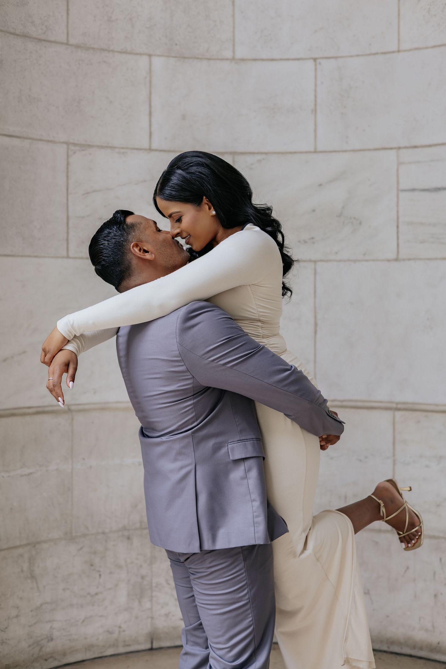 Groom lifts smiling bride against marble wall in joyful romantic embrace.