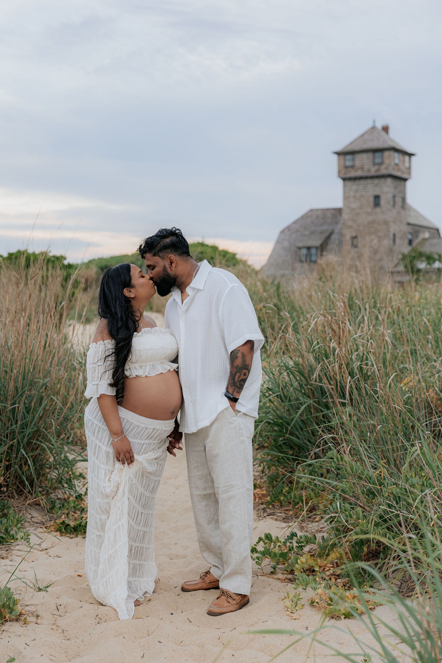 Pregnant couple in white outfits kissing on sandy beach path with tall grass and stone tower in background.