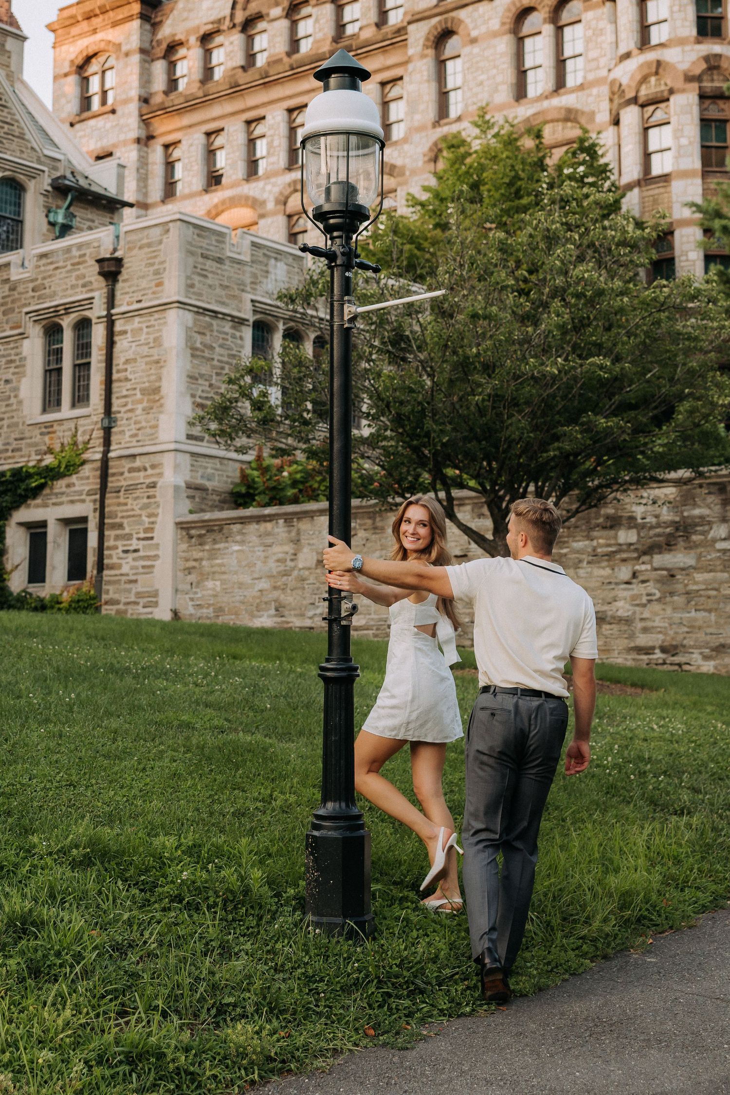 Couple dancing by a vintage street lamp on a lush green campus path beside a historic stone building.