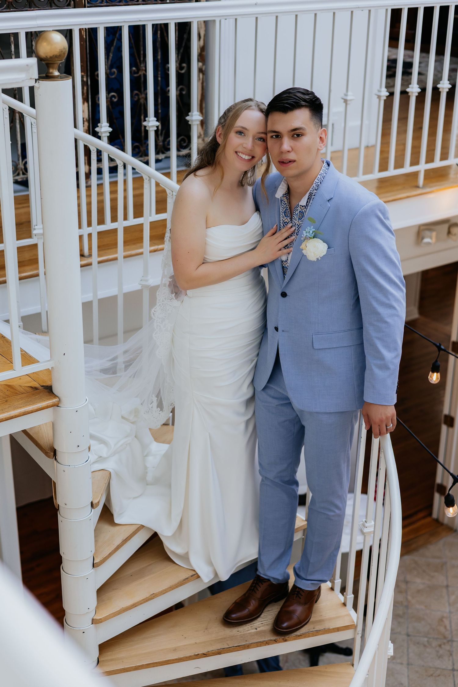 Bride and groom posing together on a white spiral staircase, she in a white gown and he in a light blue suit.