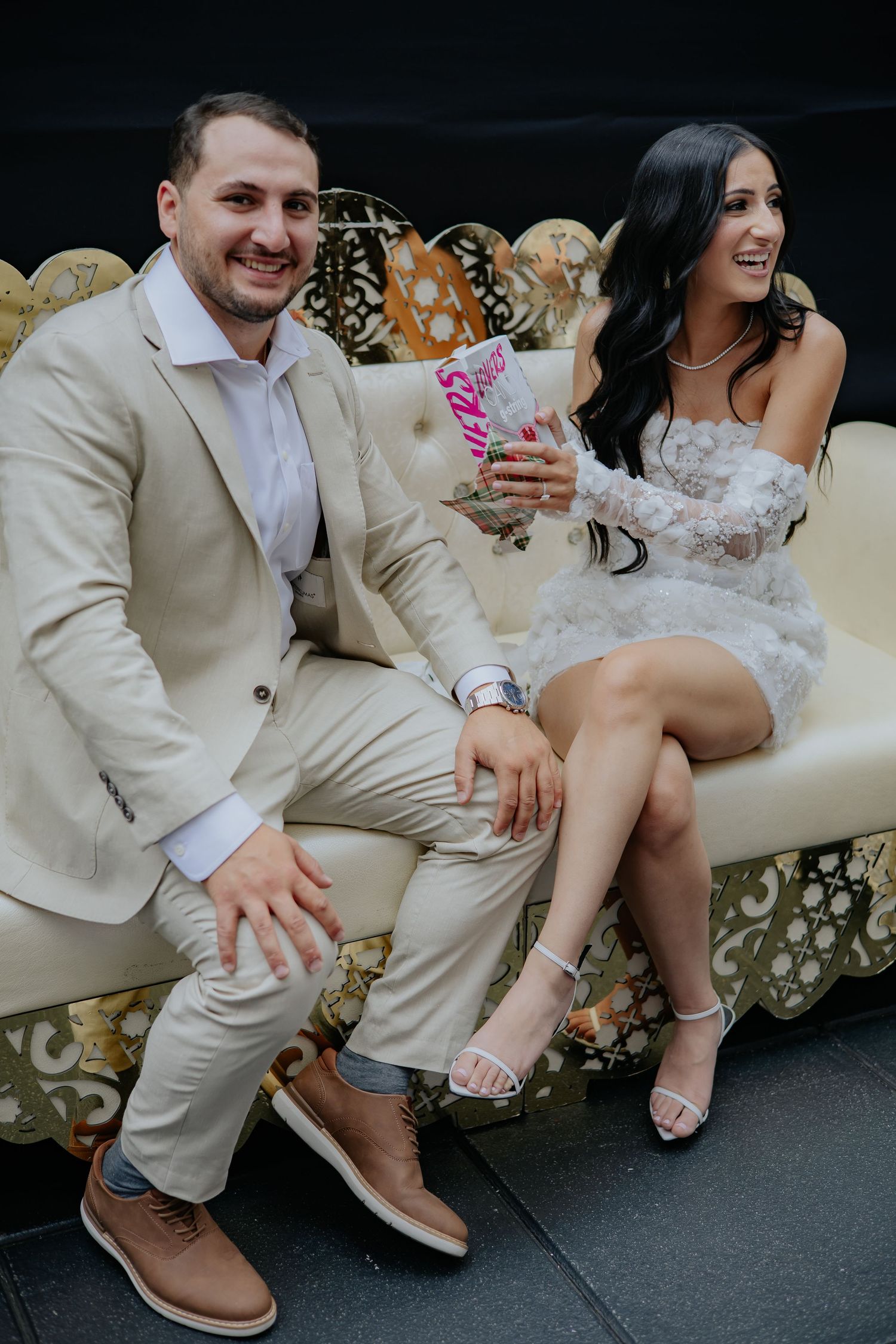 Smiling bride and groom sitting on ornate sofa holding a candy gift at wedding celebration.