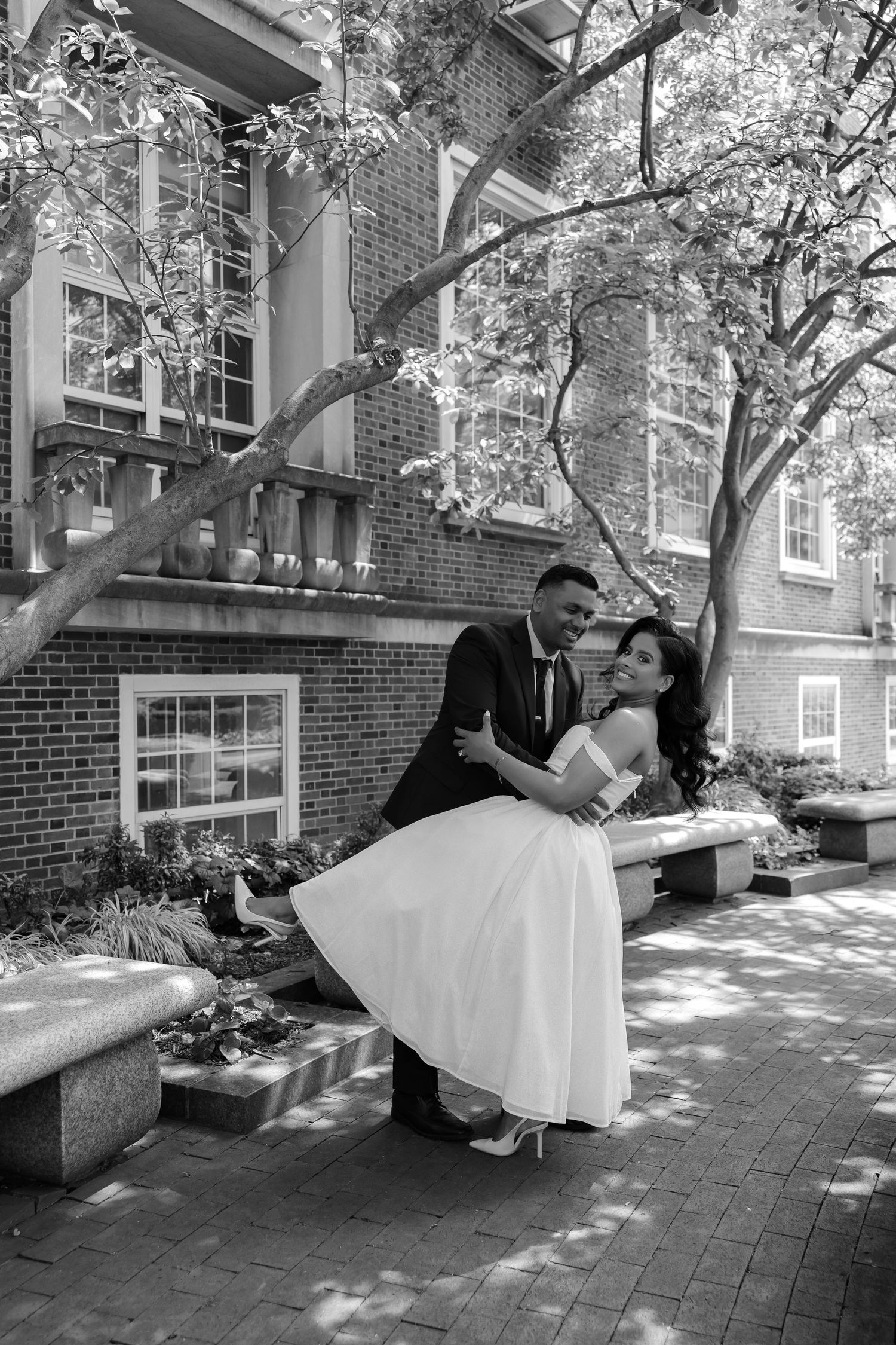 Black and white photo of a bride sitting on groom's lap on a bench in a courtyard.