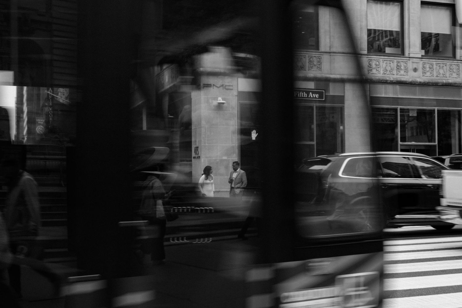 Black and white street photography of two people conversing on a city sidewalk with cars and buildings in the background.