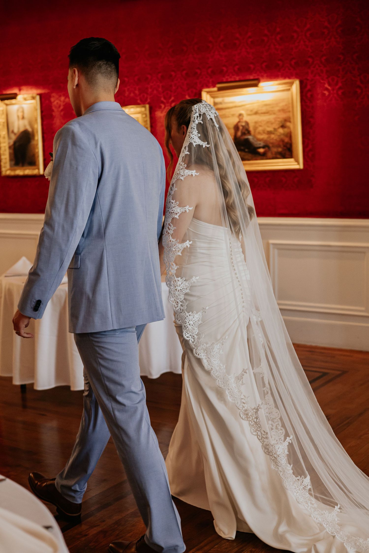 Bride and groom walking away in red-walled venue, bride in lace veil and fitted white gown.