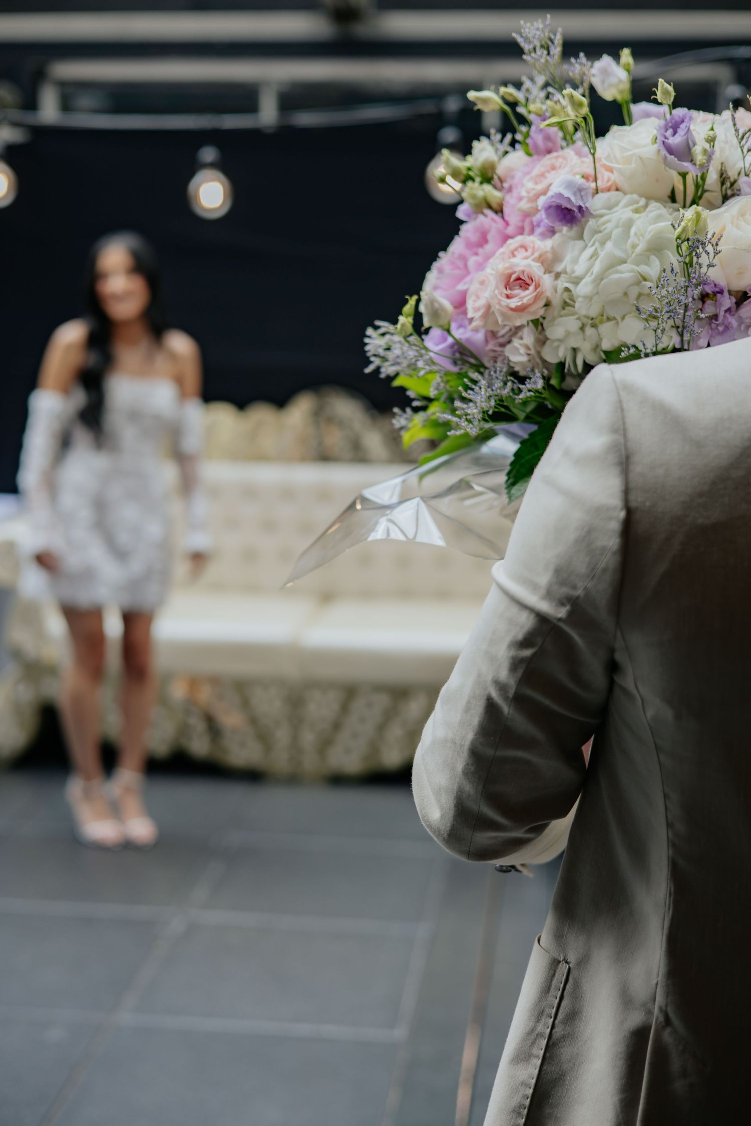 Groom holding a large floral bouquet behind his back as bride stands across the room.