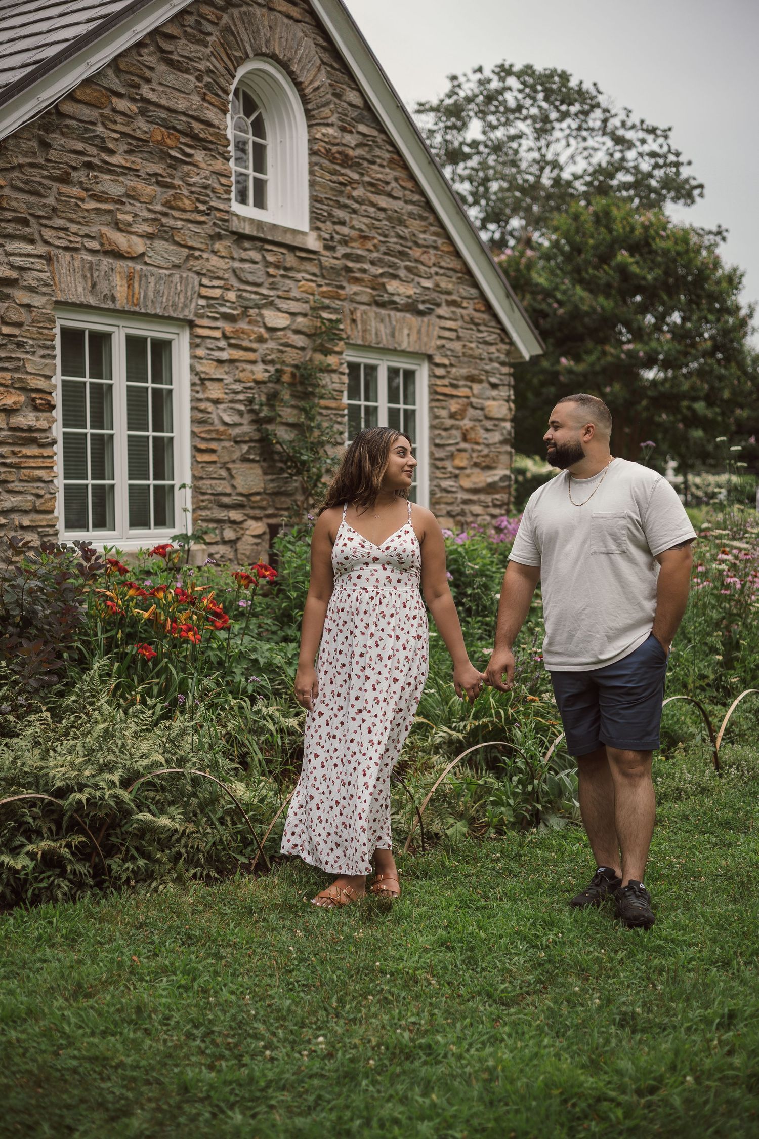 Couple holding hands walking through lush garden beside a rustic stone cottage.
