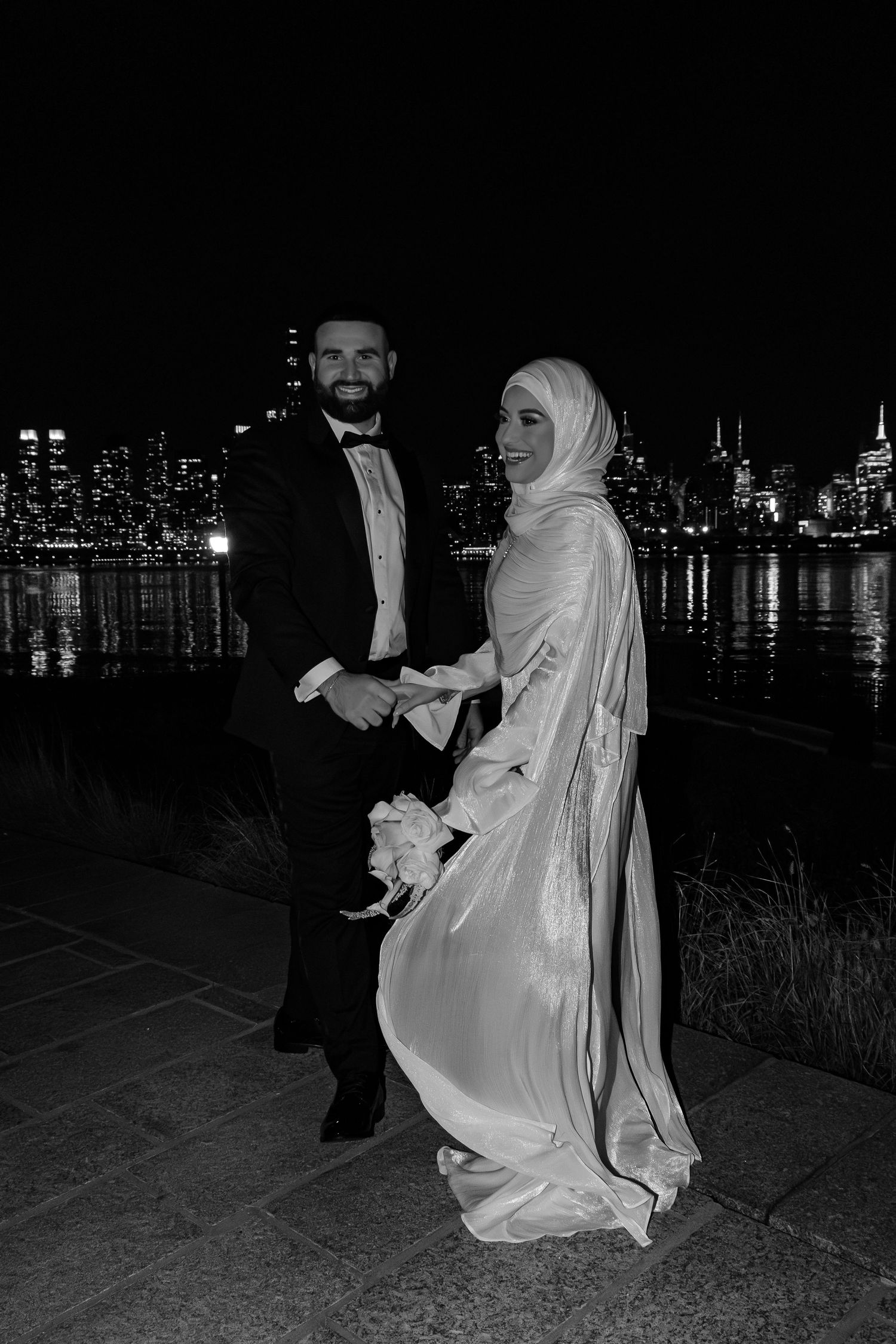 Black and white photo of Muslim wedding couple holding hands walking along waterfront at night.