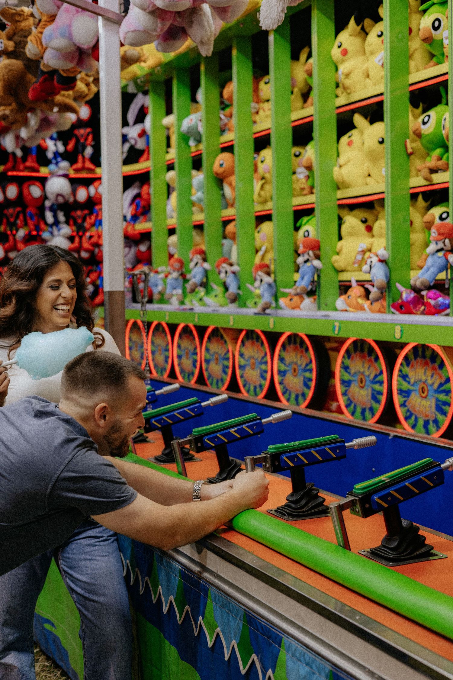 Couple laughing and playing a carnival game surrounded by colorful stuffed animal prizes at a fair.