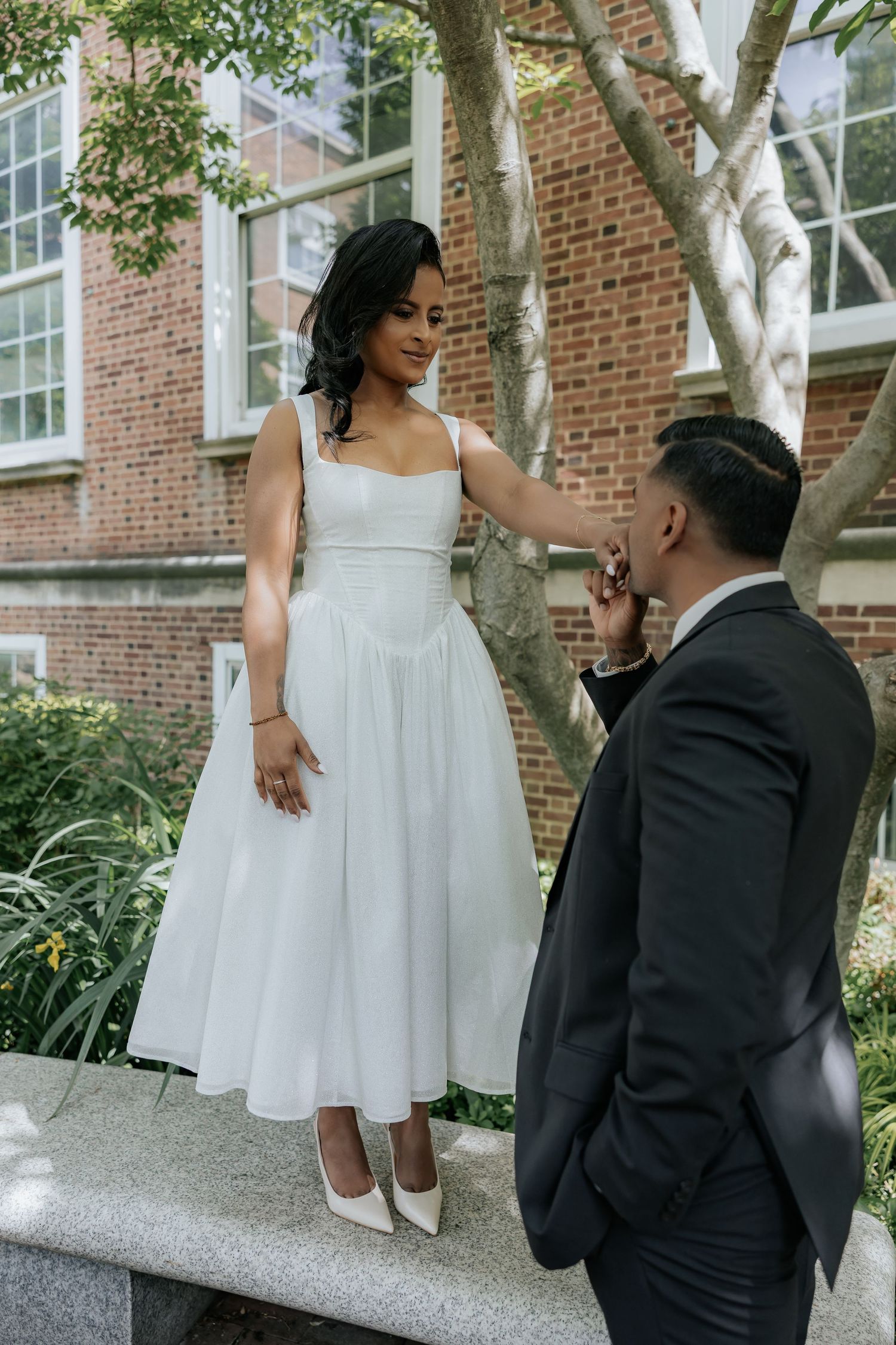 Bride standing on stone bench in white dress as groom kisses her hand in lush courtyard.