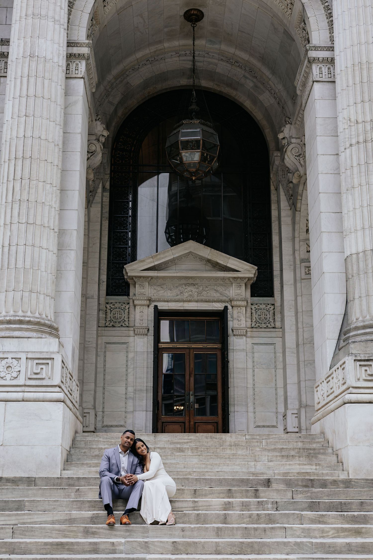 Engaged couple sitting together on grand marble steps of a neoclassical building.