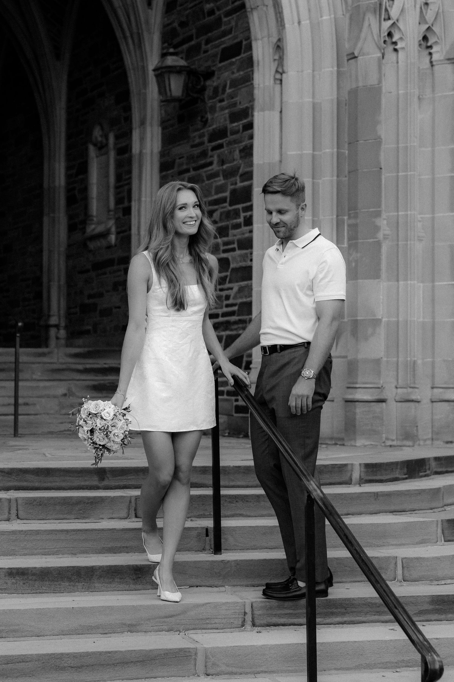 Black and white photo of a bride in a short white dress holding a bouquet with a groom on church steps.
