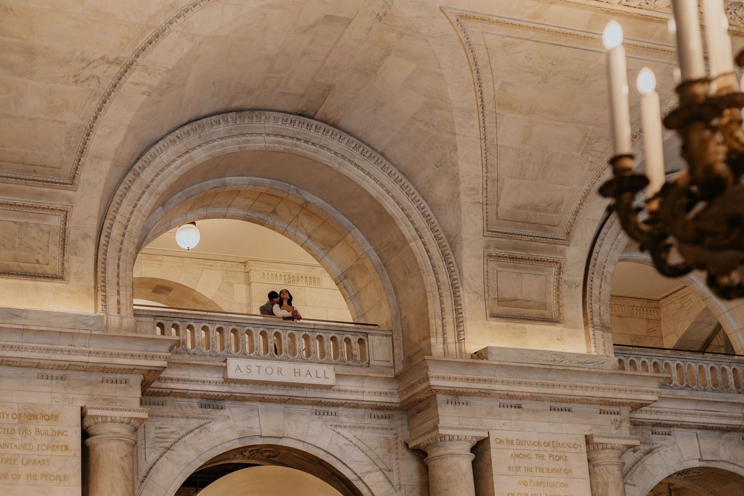 Couple embraces on upper balcony viewed through layered stone arches of grand hall.